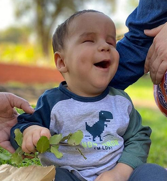 Bebé no vidente sonríe mientras sostiene una pequeña rama verde en sus manos. Lleva puesta una polera de manga larga con el dibujo de un dinosaurio azul. Está al aire libre, con fondo de césped y árboles desenfocados, acompañado por adultos que lo sostienen con ternura. La imagen transmite alegría y conexión.