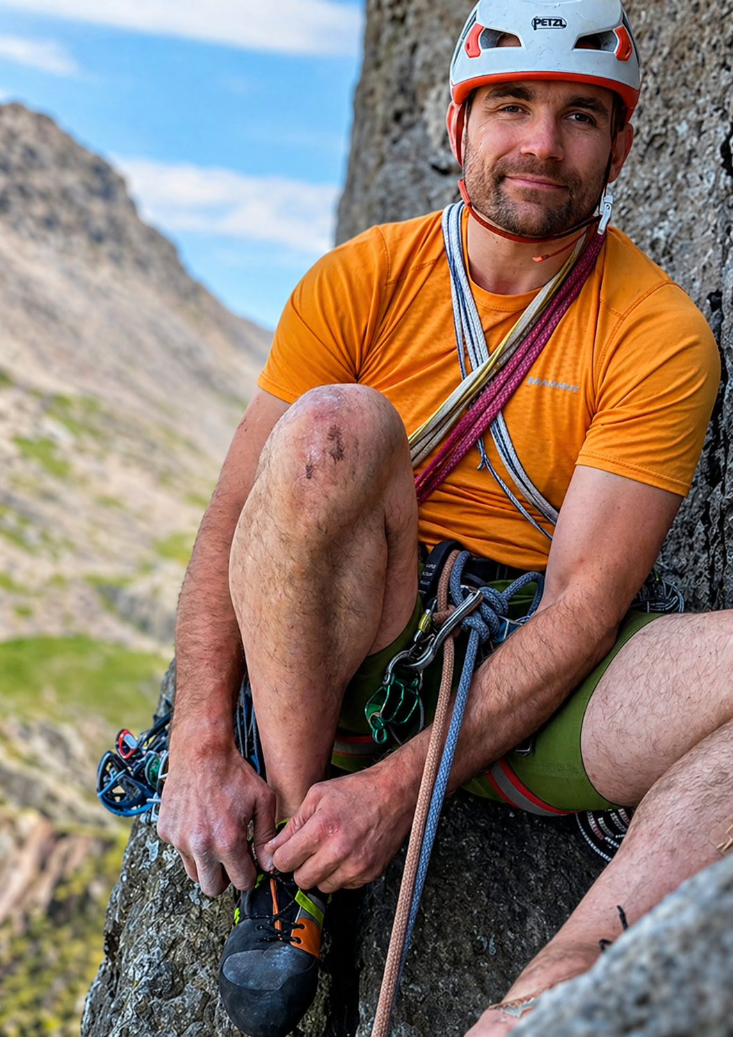 Hombre adulto de piel clara, con barba corta y expresión tranquila, sentado en una pared rocosa en plena montaña. Lleva casco de escalada blanco con detalles naranjos, polera deportiva naranja y arnés con cuerdas y mosquetones sujetos al cuerpo. Está en una pausa durante la escalada, inclinándose hacia adelante mientras amarra su zapato de escalada con ambas manos. Tiene las piernas descubiertas, con raspones y suciedad propios de la actividad. Al fondo se ve un paisaje montañoso desenfocado, que transmite altura y un entorno natural.