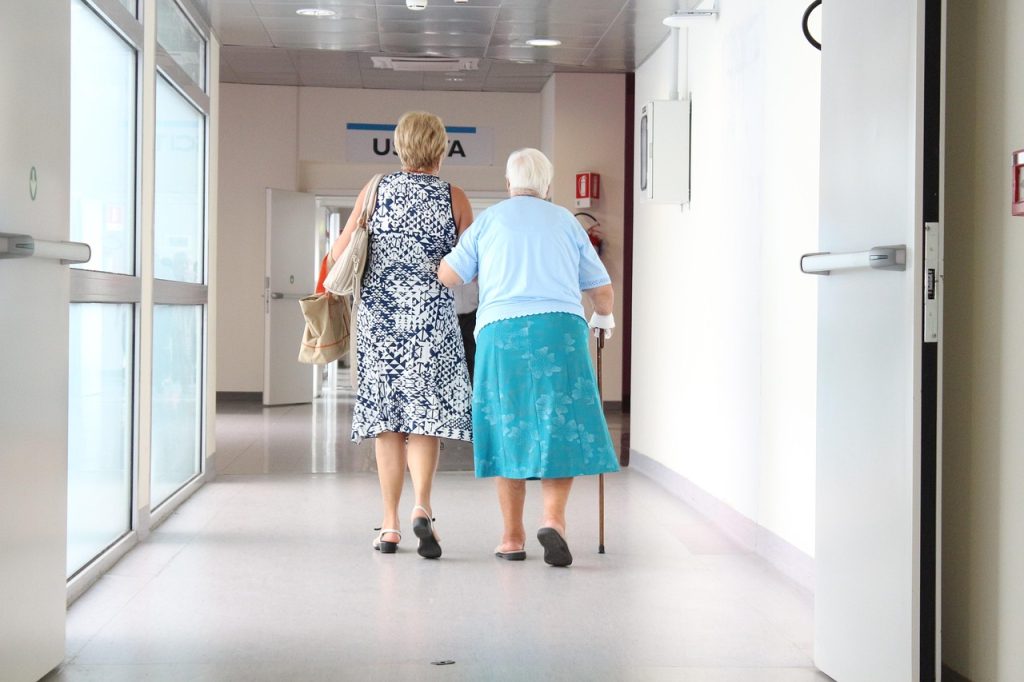 Dos mujeres mayores caminan de la mano por un pasillo. Las vemos de espalda.