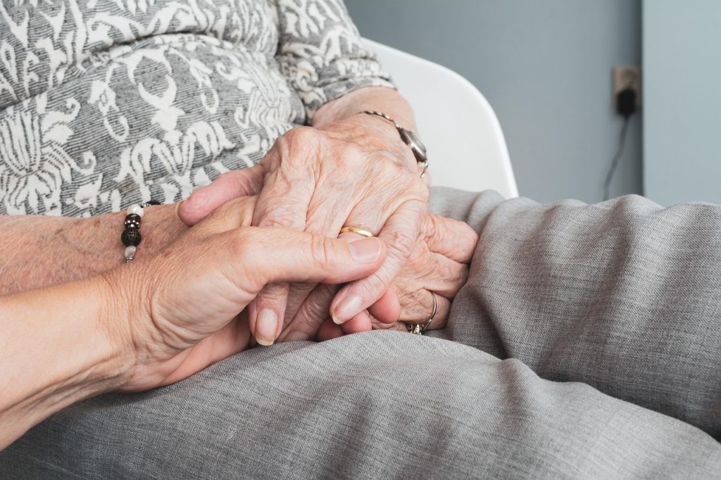 Mano de una mujer de la tercera edad sostenida por la mano de un hombre más joven. Ambas están apoyadas en el regazo de la mujer mayor.